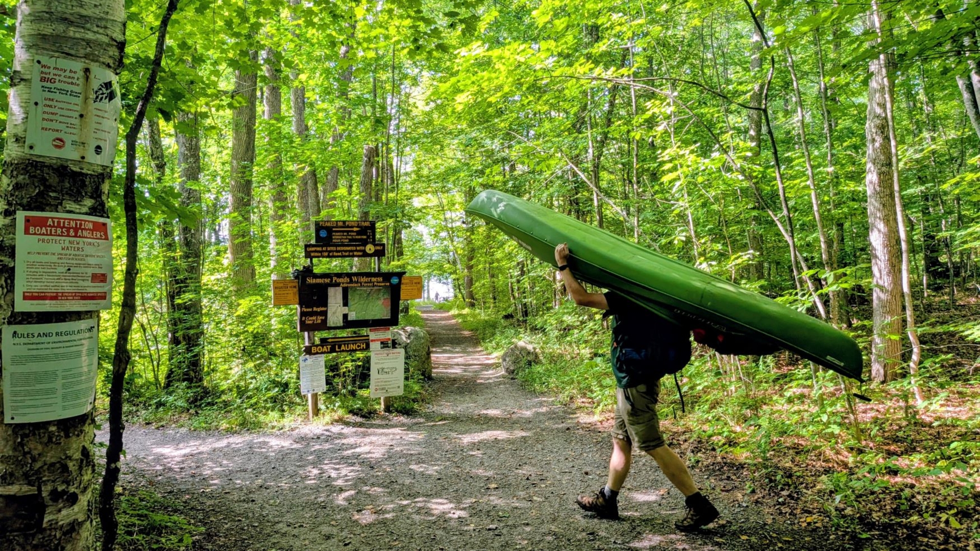 A man carries a kayak past signs pointing to 13th lake in the summer with green light filtering through the Summer canopy.